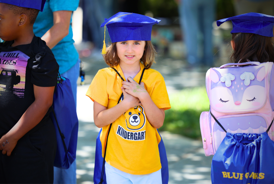 An elementary school student wearing a graduation cap smiling outside. 